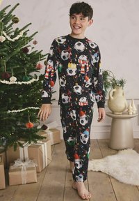 Băiat zâmbind și mergând desculț, îmbrăcat în pijamale negre de Crăciun decorate cu mingi de fotbal, aproape de un brad de Crăciun împodobit și cadouri învelite.