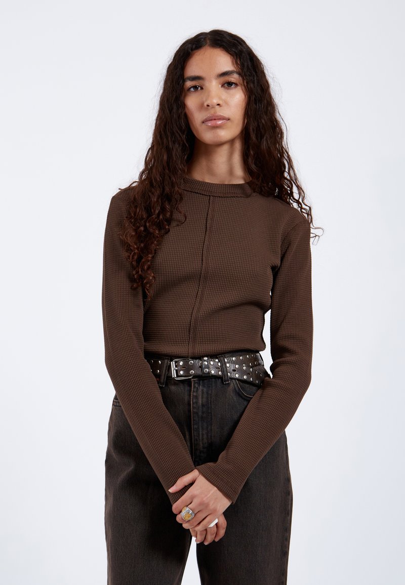 Mujer con el pelo largo y rizado que lleva una camiseta marrón de manga larga con textura, un cinturón negro con tachuelas y pantalones oscuros de talle alto, de pie con las manos entrelazadas.