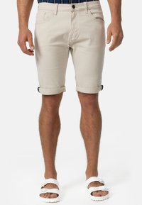 Beżowe bawełniane szorty z wywiniętym dołem, pięcioma kieszeniami i pojedynczym zapięciem na guzik, w połączeniu z białymi sandałami.