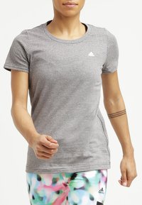 Osoba, která jde, oblečená v šedém perforovaném tričku Adidas a barevných legínách Adidas s tetováním tří čar na levém předloktí.