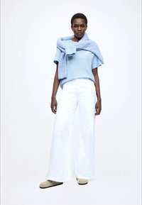 helehall sinine t-särk, valged laiade säärtega püksid ja beež jalanõud; sinine tekstuuriga kampsun heidetud üle õlgade. Minimalistlik disain, ühevärvilised riided.