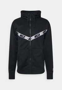 Svart zip-up hoodie med en texturerad yta, med ett diagonalt vitt Nike-logoband över bröstet och två framfickor.