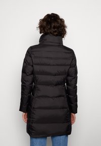 Manteau rembourré noir avec un col montant, présentant un design matelassé et des manches longues, atteignant mi-cuisse avec une texture lisse et brillante.