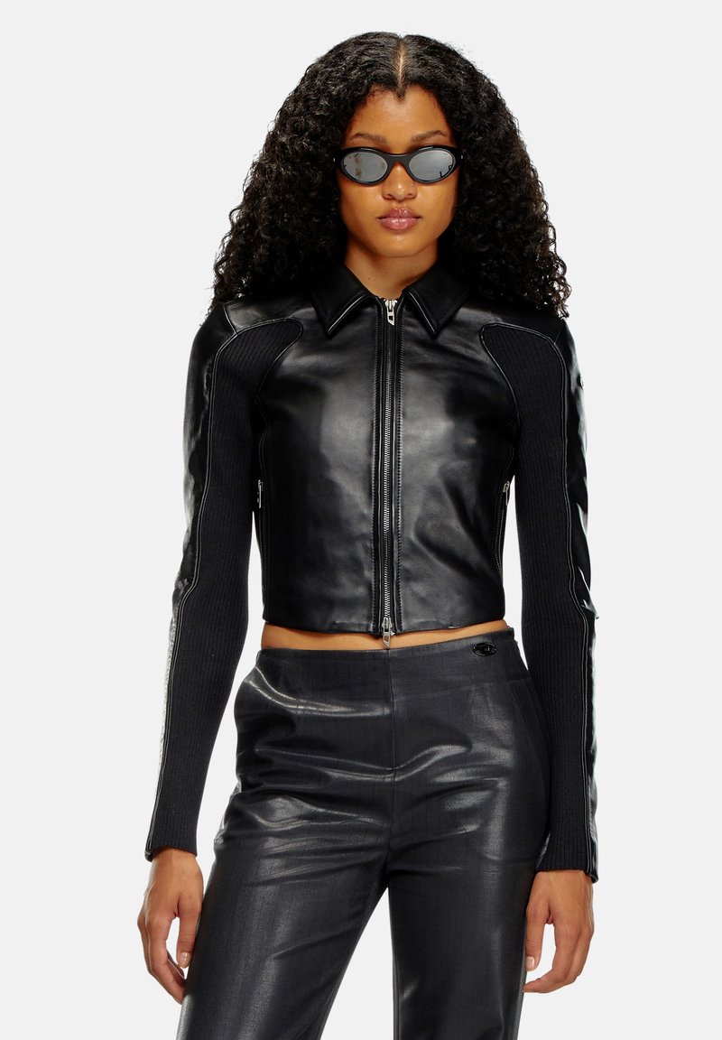 Veste en cuir noire courte avec des manches en tricot texturé, col haut et fermeture éclair à l'avant. Portée avec un pantalon en cuir noir assorti.