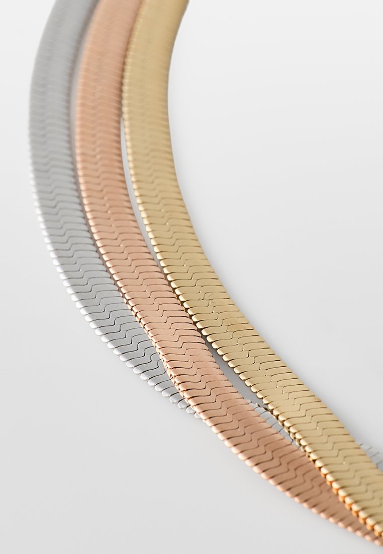 Trzy splątane metalowe łańcuchy w kolorze srebrnym, różowego złota i żółtego złota, z zygzakowatym wzorem i gładką, wypolerowaną fakturą.