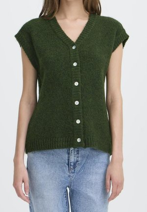 Personne portant un gilet tricoté vert foncé boutonné avec des manches courtes et un jean en denim bleu clair sur un fond uni.