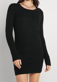 Robe-pull noire avec un design en maille côtelée texturée, col rond et manches longues. Le tissu est doux et moulant.