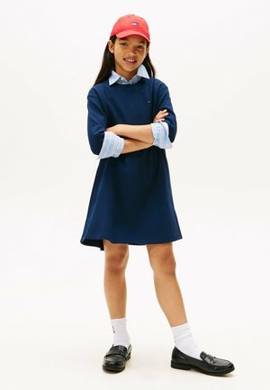 Robe marine à manches trois-quarts avec un sous-vêtement de chemise bleu clair. Chapeau rouge et mocassins noirs avec des chaussettes blanches visibles. Texture lisse.