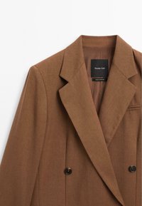 Blazer marrone doppiopetto con colletto a revers, tessuto liscio, bottoni neri e etichetta interna. Prodotto in Portogallo.