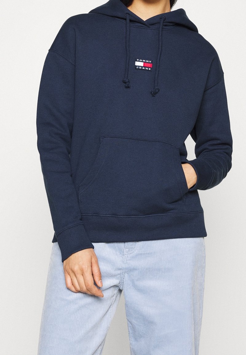Personne portant un sweat à capuche bleu marine avec une poche avant et le logo "Tommy Jeans", associé à un pantalon en velours côtelé bleu clair.