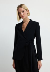 Frau mit kurzem blonden Haar, die ein schwarzes Blazer, das an der Taille gebunden ist, und einen schwarzen, gefältelten Rock vor einem einfachen, hellen Hintergrund trägt.