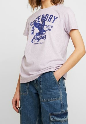 Personne portant un t-shirt violet clair avec un motif d'aigle bleu et le texte « Superdry Eagles Tokyo JPN. », associé à un jean cargo bleu ample.