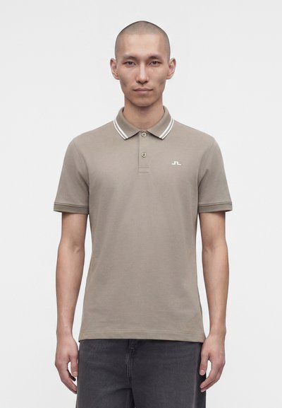 Homme à tête rasée portant un polo taupe à manches courtes avec des rayures blanches sur le col et un pantalon foncé, debout sur un fond blanc.