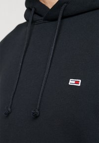 Sudadera negra con capucha y cordón ajustable, con un pequeño logo bordado en rojo, blanco y azul en el pecho. Textura de tela suave.
