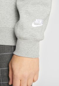 Šedá mikina se žebrovanými manžetami, na rukávu s bílým logem Nike. Viditelné detaily texturované tkaniny a přiléhavého střihu.