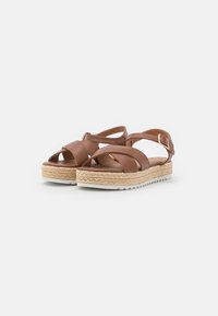 Brown leather sandals with cross straps and ankle buckle, featuring a woven jute platform and white rubber outsole.