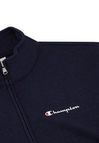 Felpa con zip di colore navy realizzata in una combinazione di cotone. Presenta un colletto alto e il logo "Champion" ricamato in bianco e rosso sul petto.