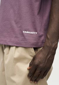 Κοντινό πλάνο σκοτεινόχρωμου μπράτσου δίπλα σε μπεζ παντελόνι και μοβ πουκάμισο Carhartt με κεντημένο όνομα της μάρκας κοντά στο τελείωμα.