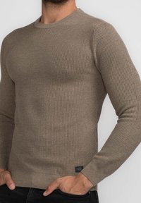 Brauner, strukturierter Strickpullover mit langen Ärmeln und Rundhalsausschnitt. Verfügt über ein kleines Logo-Etikett am Saum. Die Passform betont die Körperform.