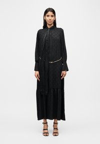 Robe noire à manches longues avec un col montant, un motif texturé et une jupe froncée. Comprend une ceinture chaîne et un détail de ruban noué.