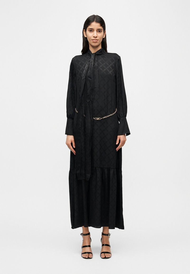 Robe noire à manches longues avec un col montant, un motif texturé et une jupe froncée. Comprend une ceinture chaîne et un détail de ruban noué.