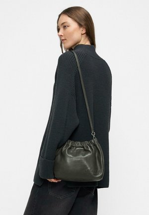 Grüne Lederbeuteltasche mit Kordelzug, glattem Finish und dezentem Branding, quer über dem Körper getragen mit einem strukturierten dunklen Pullover und schwarzen Hosen.