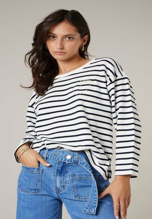 Femme aux longs cheveux foncés portant des boucles d'oreilles anneaux dorés, un haut blanc à rayures horizontales noires, et un jean taille haute bleu avec des poches avant.