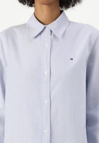 Svetlo modra in bela prugasta srajca z gumbi, ki ima klasičen ovratnik, dolge rokave in majhno izvezeno logotip na prsih.