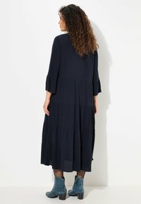 Donkerblauwe, lange jurk met gelaagd ontwerp, ronde halslijn en uitlopende mouwen. Draagt met gestructureerde blauwe enkellaarsjes.