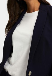 Marinblå blazer med en sned krage och en mässingsknapp. Bärs över en vit skjorta. Blazern har en slät yta och skräddarsydd design.