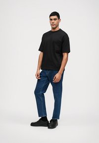 Μαύρο βαμβακερό T-shirt κοντό μανίκι με τσέπη στο στήθος, συνδυασμένο με μπλε τζιν παντελόνι και μαύρα αθλητικά, επιδεικνύοντας μια χαλαρή εφαρμογή.