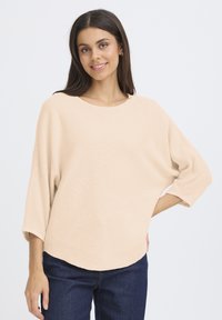 Beżowy sweter na drutach z szerokim, okrągłym dekoltem, r�ękawami o długości do łokcia i luźnym krojem, zestawiony z ciemnoniebieskimi dżinsami.