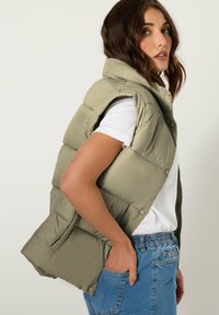 Zielono-oliwkowa kamizelka z ociepleniem, z wysokim kołnierzem, zapięciem na guziki i bocznymi kieszeniami. Nosi się ją na białym T-shircie i dżinsowych szortach. Gładka tekstura.