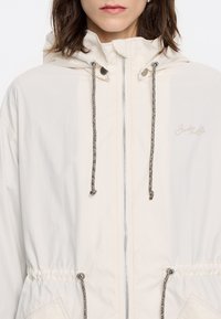 Leichte cremfarbene Jacke mit Kapuze, Kordelzug in der Taille und silbernem Reißverschluss. Besondere Merkmale sind die schwarz-weißen gemusterten Kordelzüge und das Logodetail.