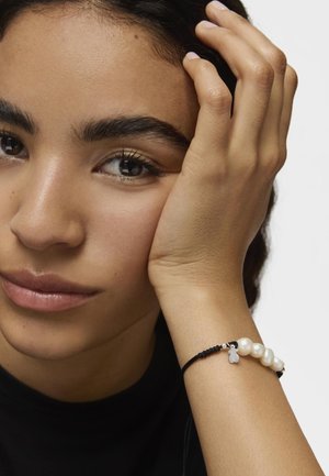Bracciale intrecciato nero con perle bianche e un piccolo ciondolo. Inquadratura ravvicinata di una mano appoggiata sul viso con pelle liscia e trucco naturale.