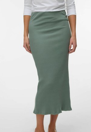 Kvinde iført en ribbet, salviegrøn, midilængde nederdel med høj talje kombineret med en hvid långærmet top, vist fra talje til fødder.
