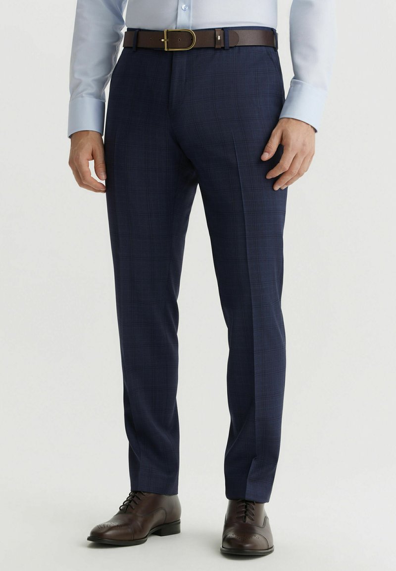 Granatowe spodnie dopasowane, z delikatnym wzorem w kratkę, teksturą materiału oraz brązowym skórzanym paskiem. Całość uzupełniają brązowe buty eleganckie.