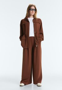 Woman standing wearing oversized brown jacket with large pockets, matching wide-leg pants, white shirt, beige shoes, and black sunglasses.