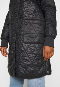 Cappotto trapuntato nero con una texture liscia e lucida. Presenta polsini a coste, una tasca frontale e chiusure a scatto lungo la patta.