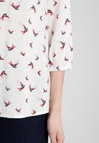 White blouse with a bird pattern in red, blue, and black. Features three-quarter sleeves and elastic cuffs. Soft fabric texture.