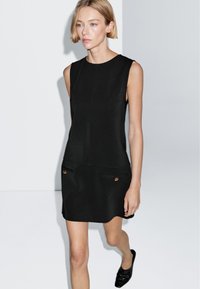 Schwarzes ärmelloses Kleid mit geradem Schnitt, das zwei Vordertaschen mit goldenen Knöpfen aufweist. Glattes Material mit minimaler Textur.