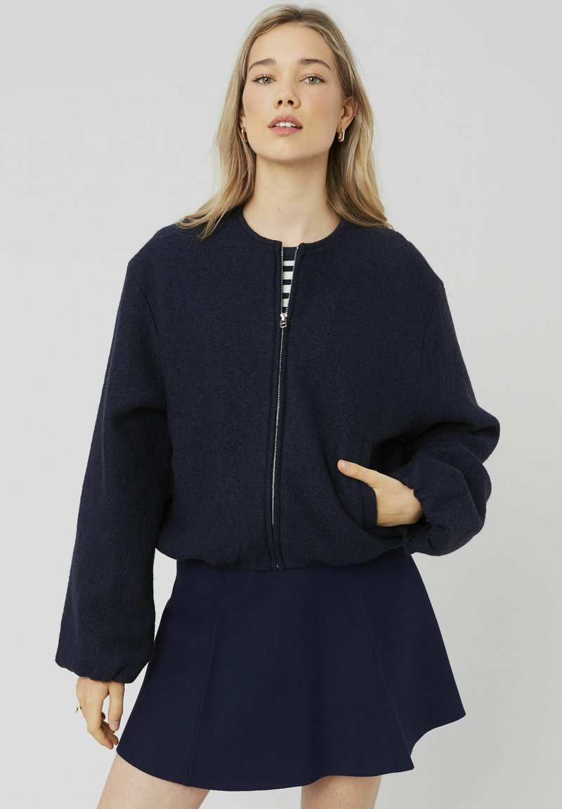 Blouson aviateur bleu marine en tissu doux et texturé, avec une fermeture éclair sur le devant, un col rond et des manches à poignets. Porté par-dessus un haut rayé.