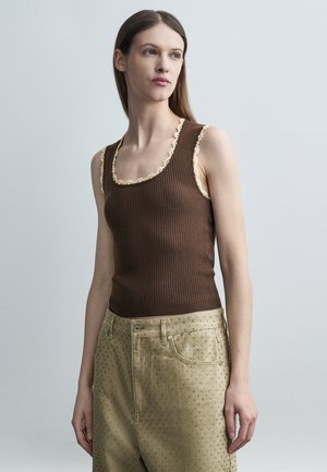 Žena s dlhými hnedými vlasmi, oblečená v priliehavom hnedom rebrovanom tielku s čipkovaným lemom a béžových nohaviciach s vysokým pásom a malým bodkovaným vzorom, hľadí doprava.