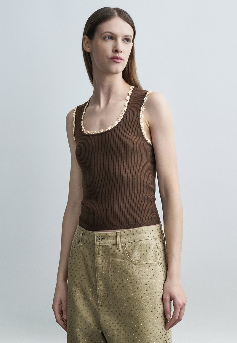 Γυναίκα με μακριά καστανά μαλλιά, φορώντας στενό καστανό ριμπ τοπ με δαντελένιο τελείωμα και μπεζ παντελόνι ψηλόμεσο με μικρό πουά μοτίβο, κοιτάζοντας προς τα δεξιά.