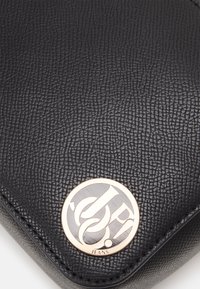 Czarna skórzana torebka o fakturowanej powierzchni, ozdobiona złotym metalowym logo z okrągłym wzorem i detalami wzoru na powierzchni.