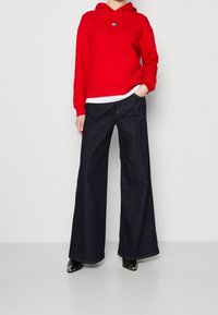 Czerwona bawełniana bluza z kapturem z przednią kieszenią i logo, w połączeniu z szerokimi ciemnoniebieskimi dżinsami i czarnymi botkami na obcasie, na jasnoszarym tle.