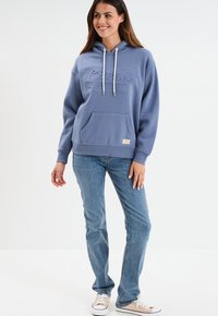 Felpa blu con cappuccio, tasca frontale, dettagli con cordino e logo impresso. Abbinata a jeans dritti in lavaggio chiaro e sneakers.