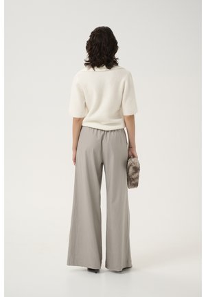 Kobieta z krótkimi kręconymi włosami, ubrana w biały sweter z krótkim rękawem i szerokie beżowe spodnie, trzymająca małą pluszową kopertówkę, stoi tyłem do kamery.