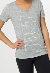 Camiseta de algodón gris con diseño de cuello en V, que presenta grandes letras en bloque blancas que dicen "JUST" en la parte delantera. Mangas cortas, forma ajustada.