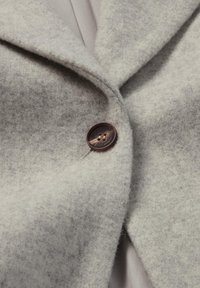 Graue Wolljacke mit strukturierter Oberfläche, versehen mit einem glatten, braunen Knopf und einem Revers mit Kerbe.